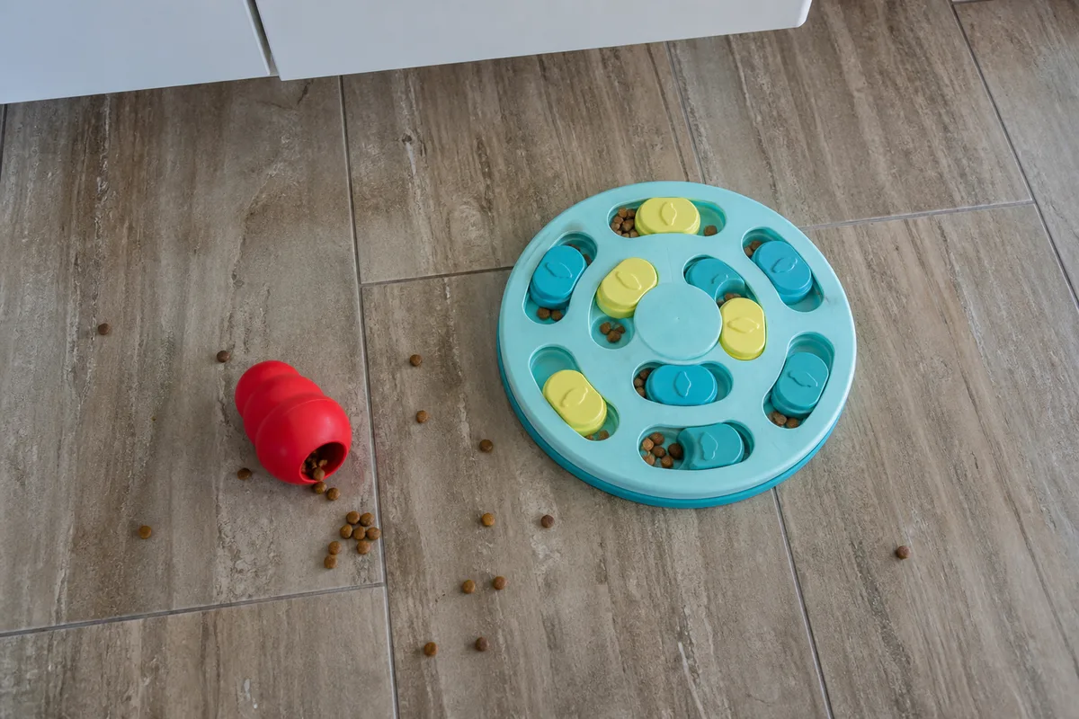 A rubber food-stuffed toy and a puzzle feeder on a kitchen floor
